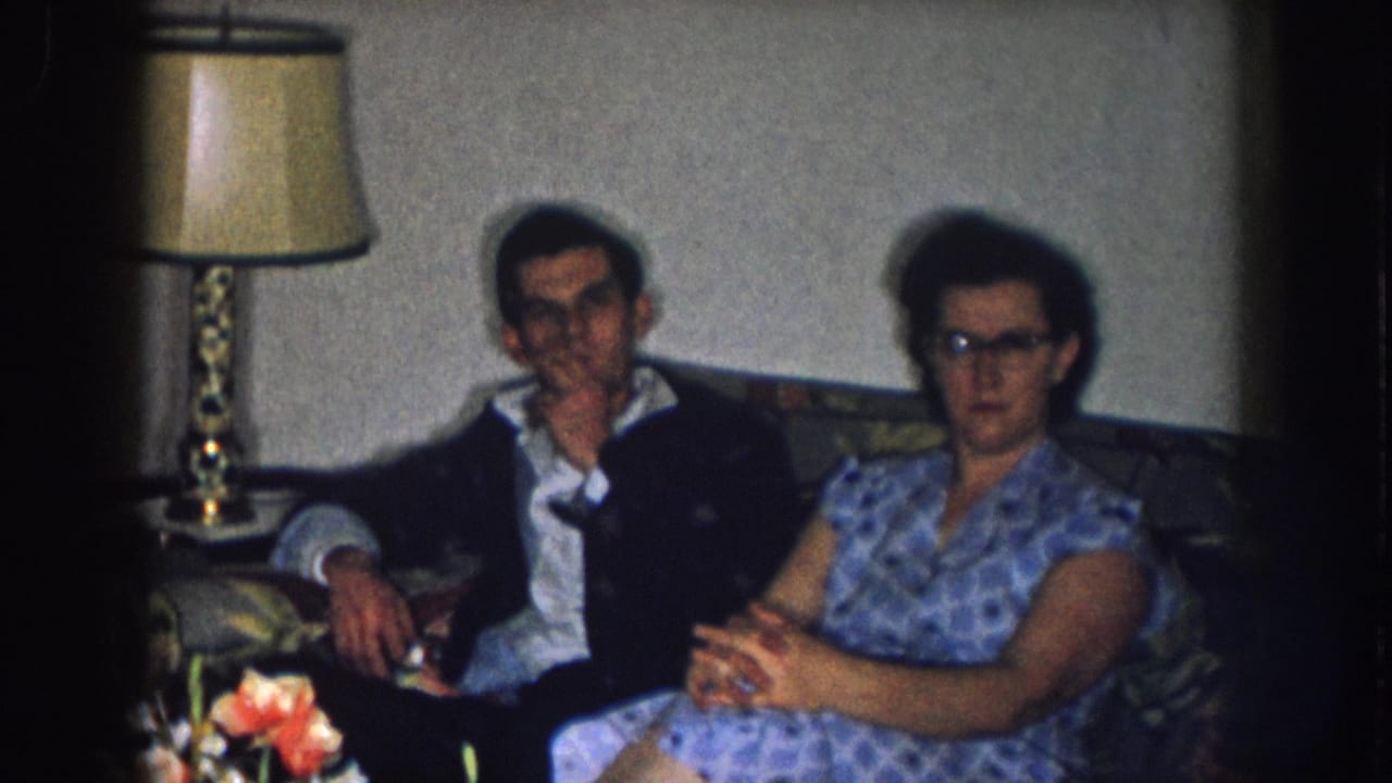 1957 Nebraska 8mm home movie — man in dark jacket and woman in blue-print dress with cat-eye glasses on a patterned sofa, ornate lamp and floral arrangement at left.