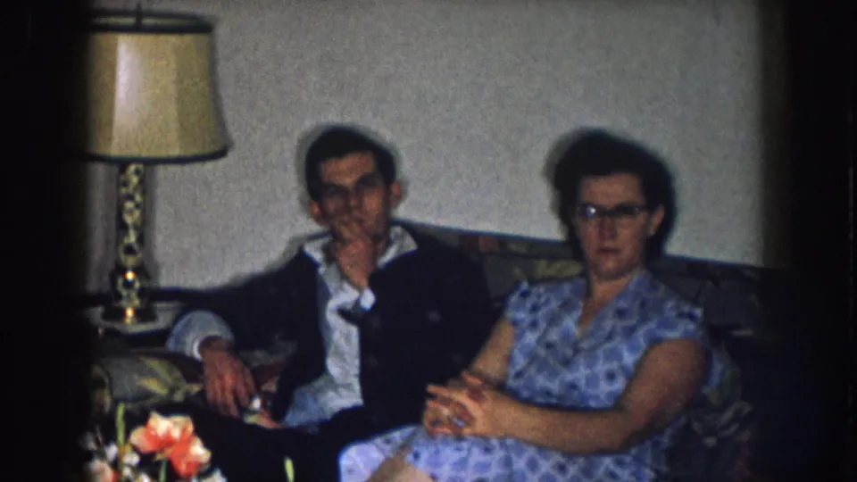 1957 Nebraska 8mm home movie — man in dark jacket and woman in blue-print dress with cat-eye glasses on a patterned sofa, ornate lamp and floral arrangement at left.
