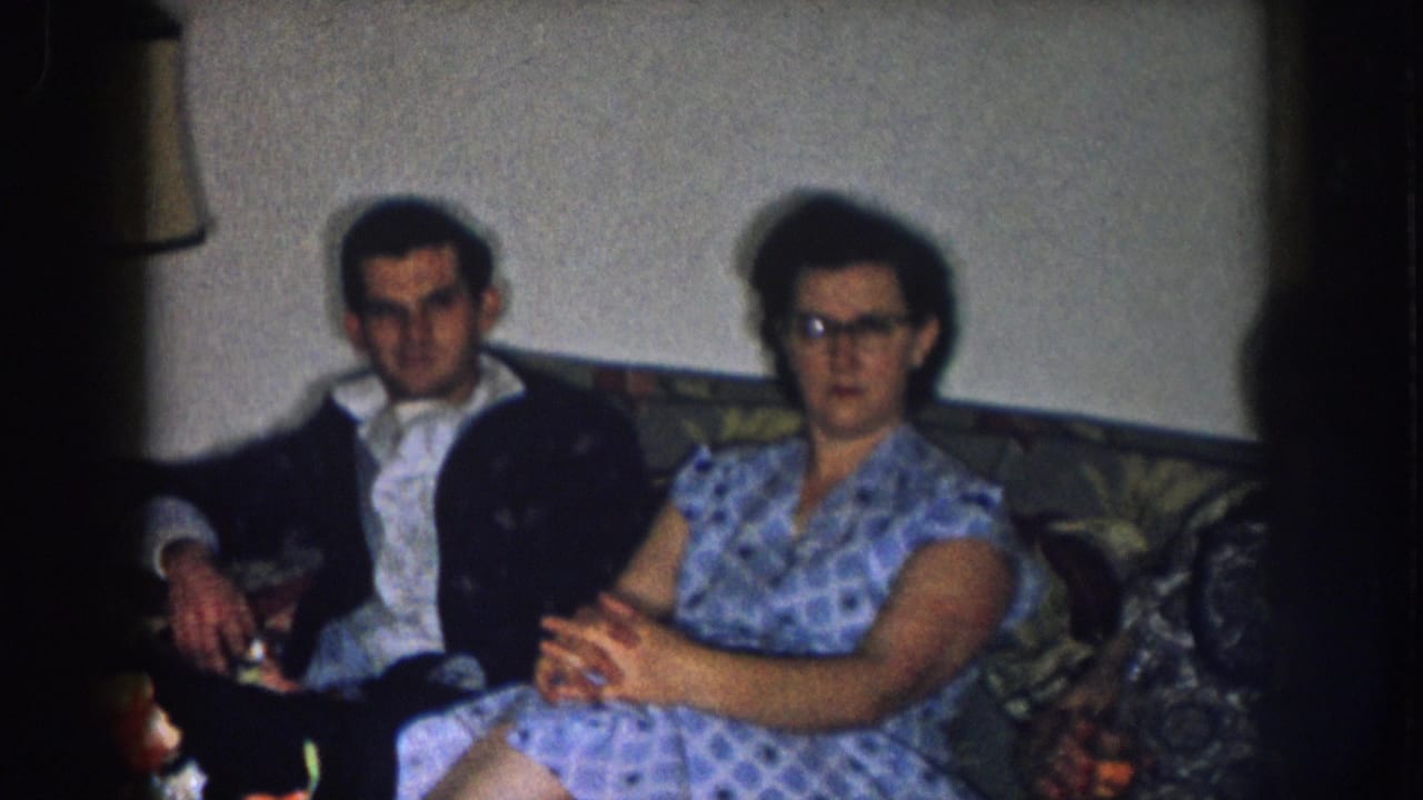 Man and woman remain on sofa; woman's cat-eye glasses more distinct, film-edge vignette visible at right, orange flowers at lower right.