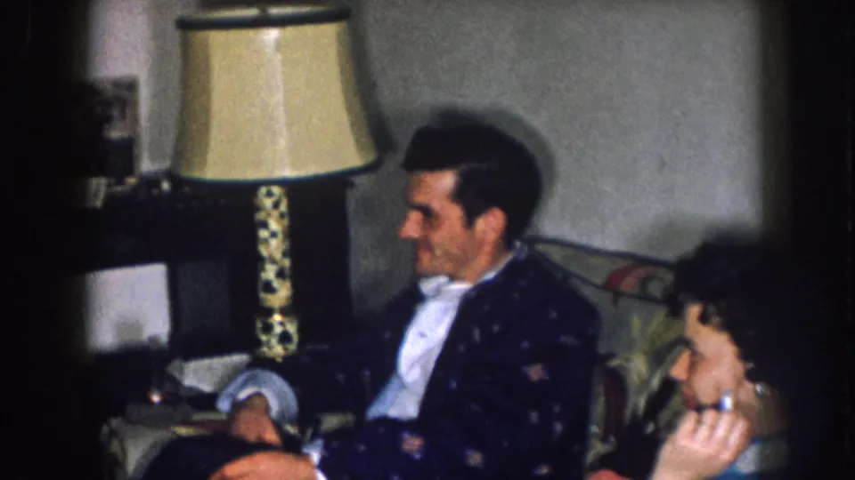 1957 Nebraska 8mm home movie — camera reframe shows man in dark jacket in closer view, ornate lamp prominent at left; second seated figure appears at lower right.