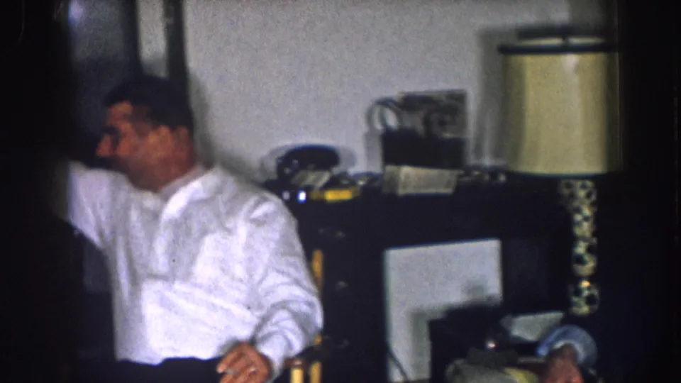 1957 Nebraska 8mm home movie — man in white shirt gestures or reaches toward right; table lamp and dark rectangular object at frame right; candid indoor scene.
