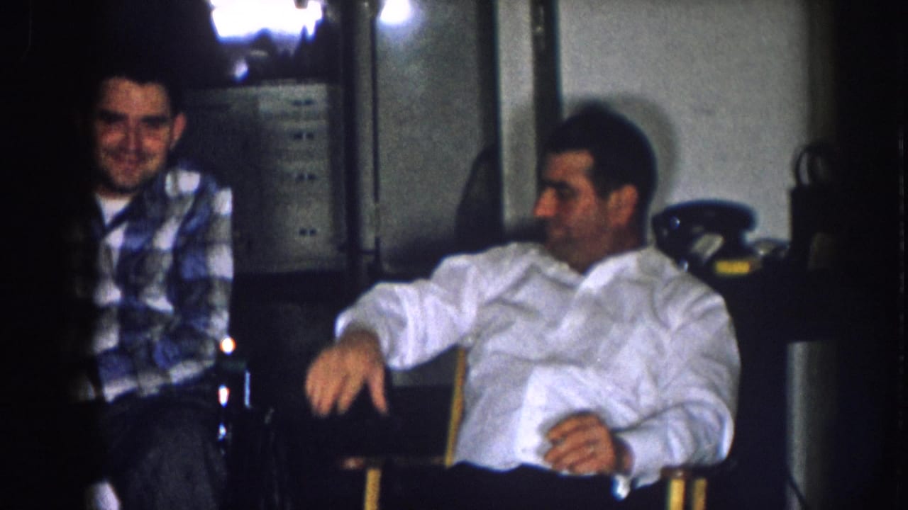 Man in white button-down shirt and man in blue plaid shirt seated together in a dimly lit 1950s domestic interior, Nebraska, c. 1957.