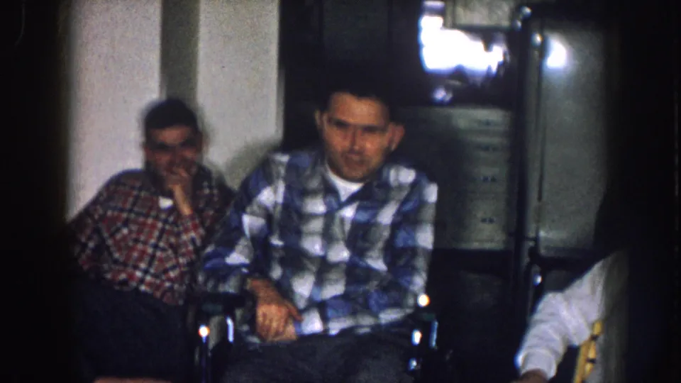 Two men in red and blue plaid shirts in a 1950s home interior, Nebraska c. 1957; an early boxy television set is visible in the background.