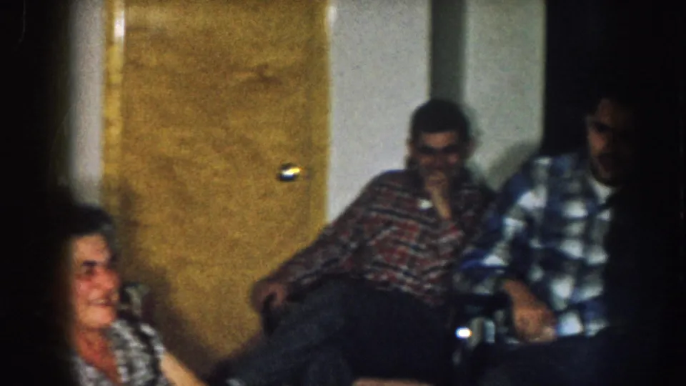 Older woman and two men in plaid shirts in a 1950s home interior, Nebraska c. 1957; a wood-paneled interior door is visible behind them.