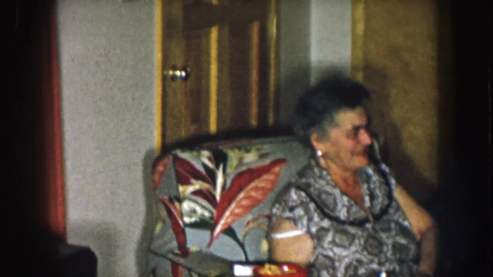 Older woman in a patterned dress holding a white drink cup, seated in a distinctive tropical-leaf floral armchair in a 1950s home interior, Nebraska c. 1957.