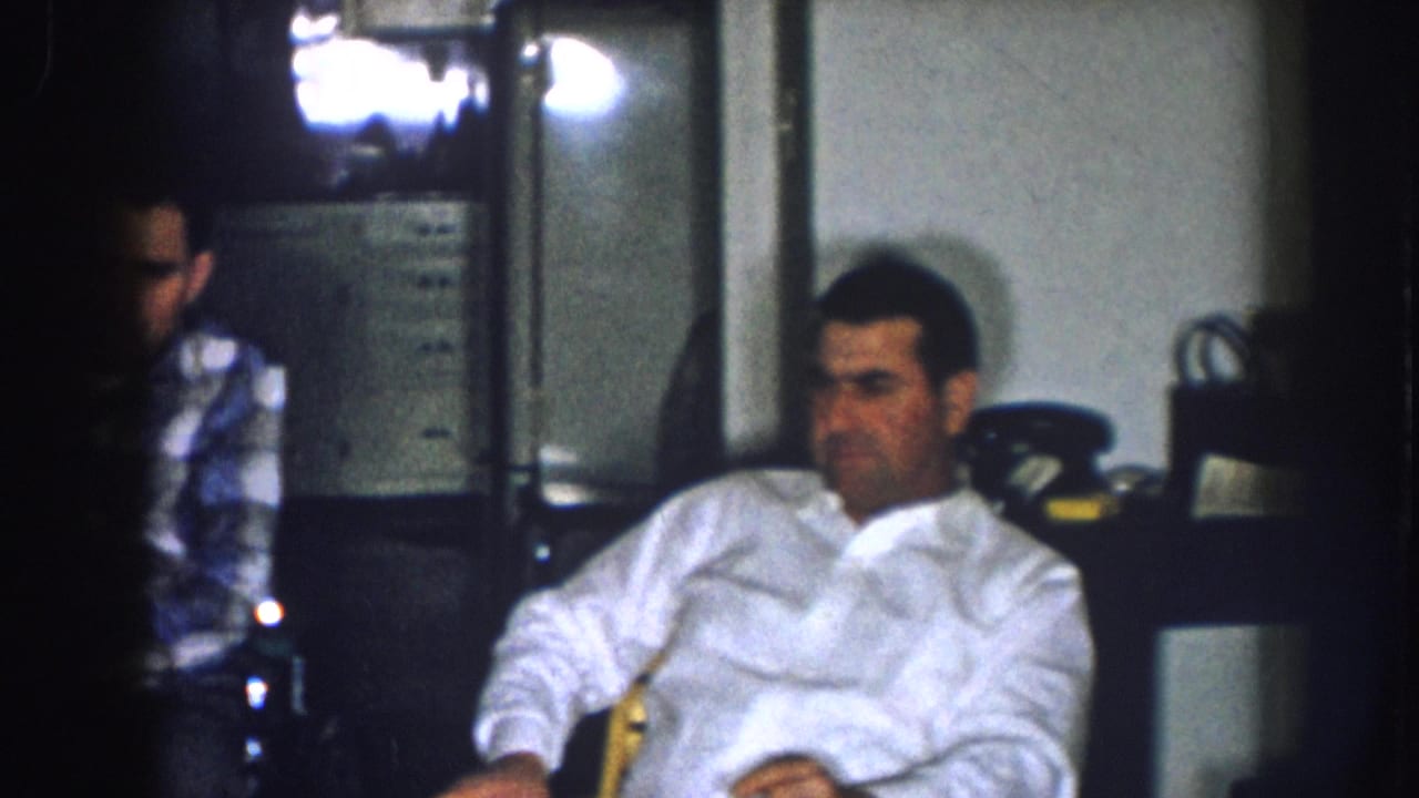 Man in white button-down shirt relaxes in an armchair in a dimly lit interior; partial figure in plaid shirt at frame edge; consistent with 1957 Nebraska 8mm home-movie footage.