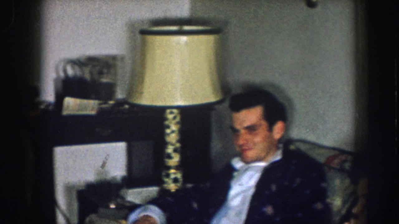 Man in dark blazer seated in a living room chair, with a period cream-shaded table lamp and dark wood furniture behind him; consistent with a late-1950s domestic interior in Nebraska.