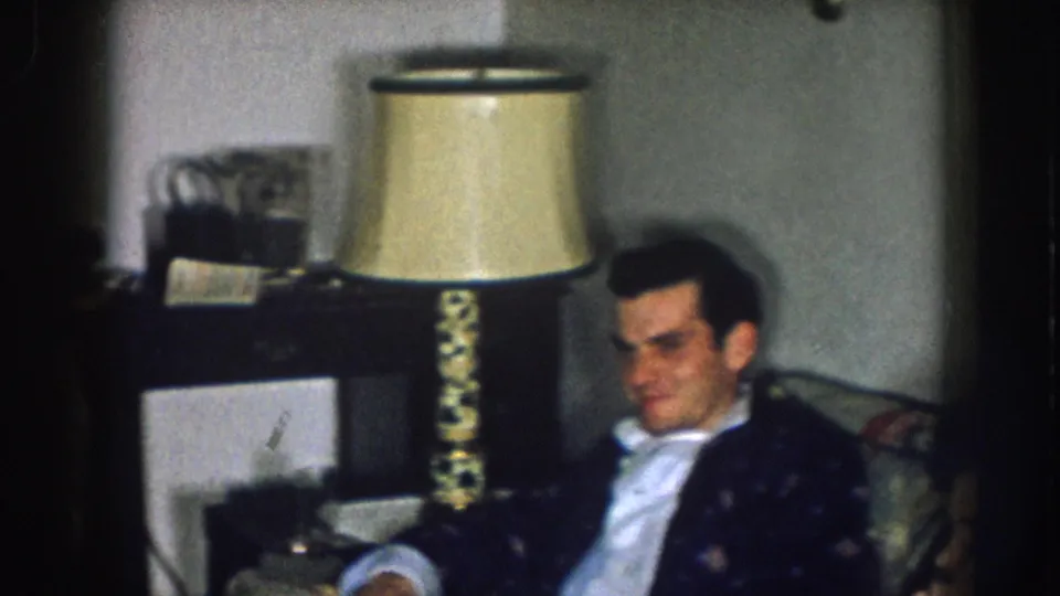 Man in dark blazer seated in a living room chair, with a period cream-shaded table lamp and dark wood furniture behind him; consistent with a late-1950s domestic interior in Nebraska.