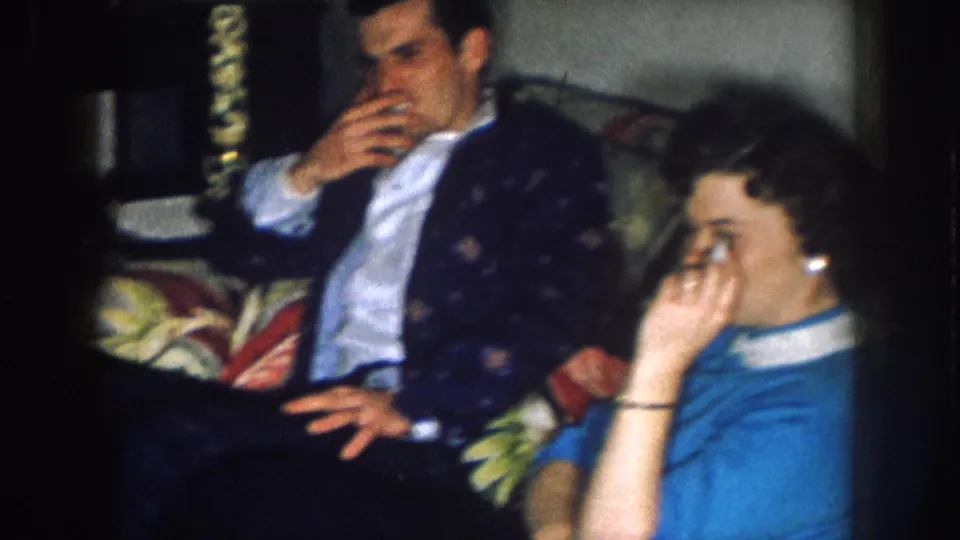 Man in dark patterned sport coat and woman in blue top sit on a floral-upholstered sofa smoking cigarettes; candid domestic gathering consistent with 1950s 8mm home-movie footage.