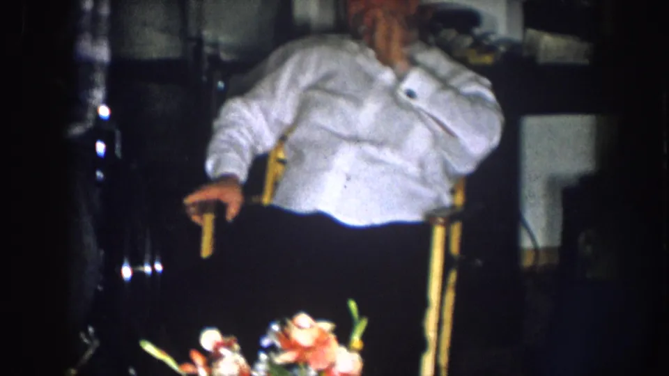 Man in white shirt relaxes in a yellow-framed wooden chair in a dim living room, with a colorful floral arrangement in the foreground; consistent with a late-1950s domestic interior.