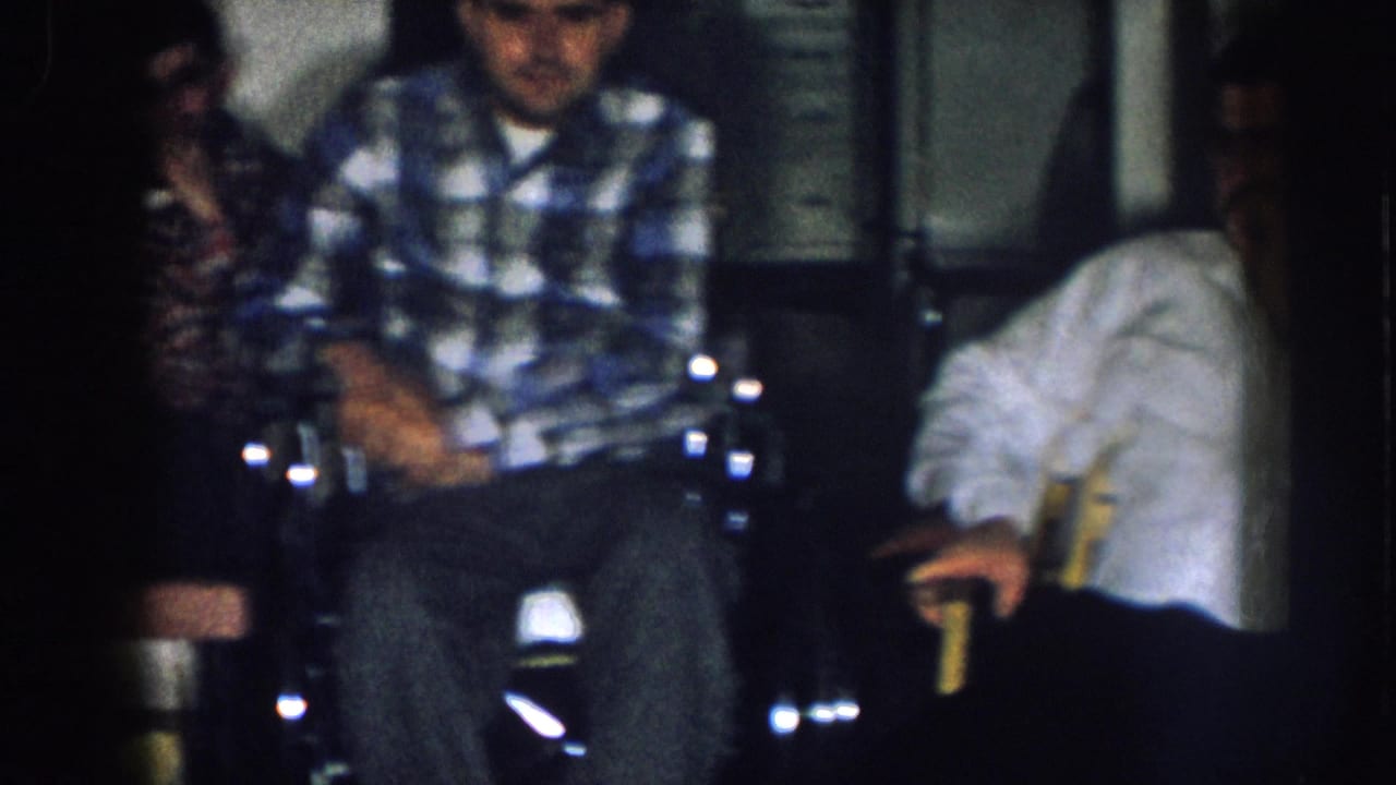 Indoor family gathering circa 1957 Nebraska; man in blue plaid shirt in wheelchair with others nearby in a home interior.