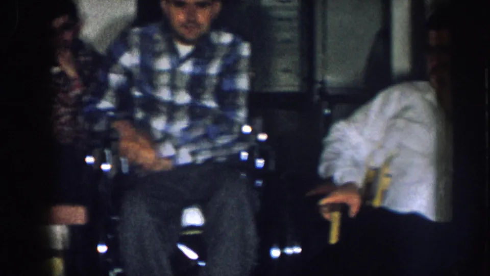 Indoor family gathering circa 1957 Nebraska; man in blue plaid shirt in wheelchair with others nearby in a home interior.