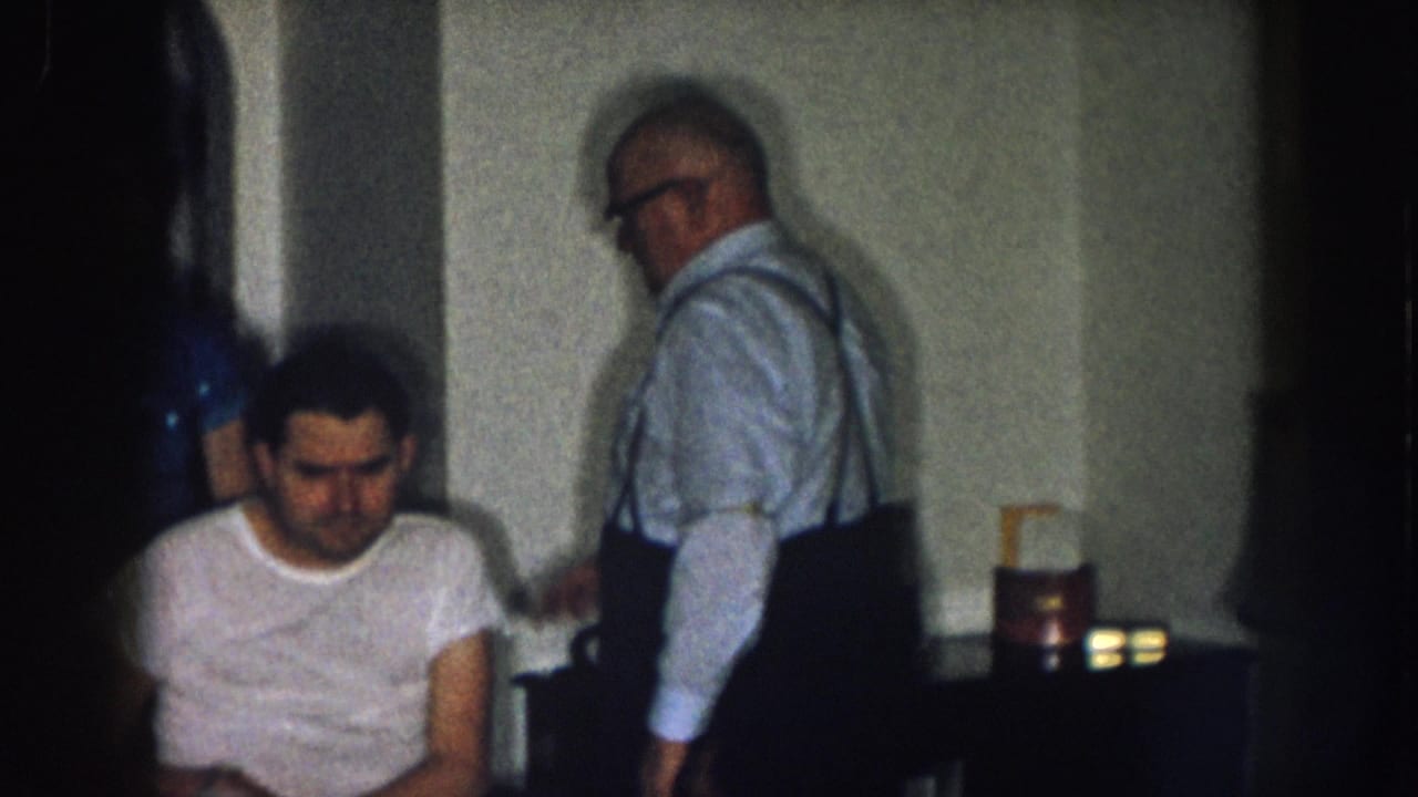 Older man in light blue shirt with suspenders and glasses stands beside a younger man in white t-shirt seated in a wheelchair, indoor setting.