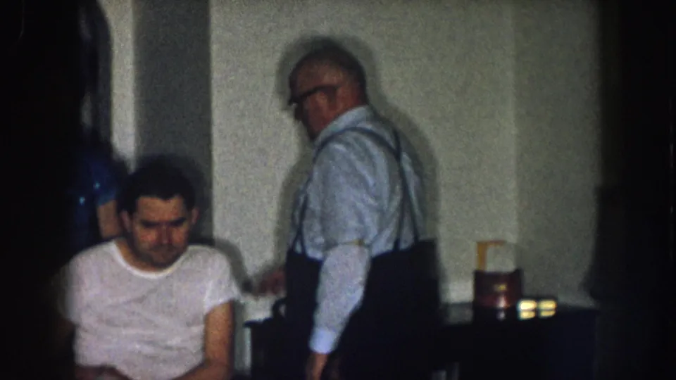 Two men in a home interior circa 1957 Nebraska — older man with suspenders and glasses beside a younger man in white t-shirt in a wheelchair.