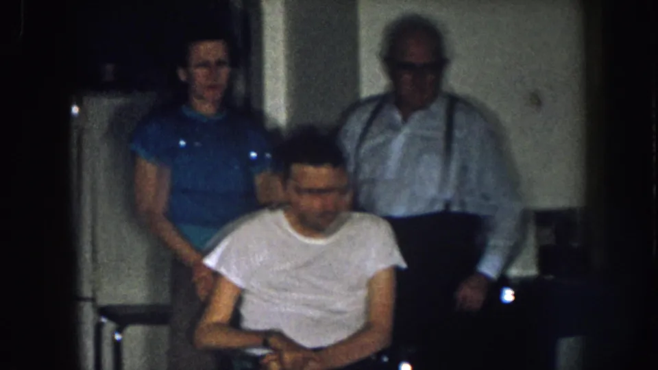 Three men at a domestic gathering circa 1957 Nebraska; man in wheelchair in foreground, older man with suspenders and younger man in blue t-shirt behind.