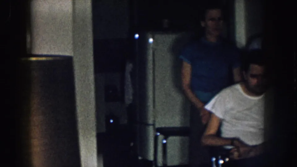 Man in white t-shirt in wheelchair at a home interior gathering circa 1957 Nebraska, with another man in blue t-shirt partially visible at left.