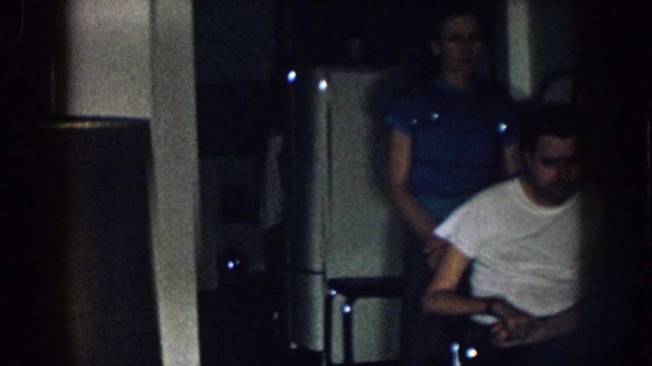 Young adult man in wheelchair with woman in blue short-sleeved shirt standing behind him, low-light kitchen interior, Nebraska USA c. 1957 8mm home movie.