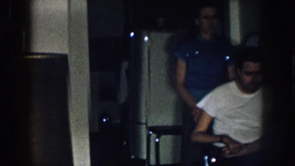 Young adult man in wheelchair with woman in blue short-sleeved shirt standing behind him, low-light kitchen interior, Nebraska USA c. 1957 8mm home movie.
