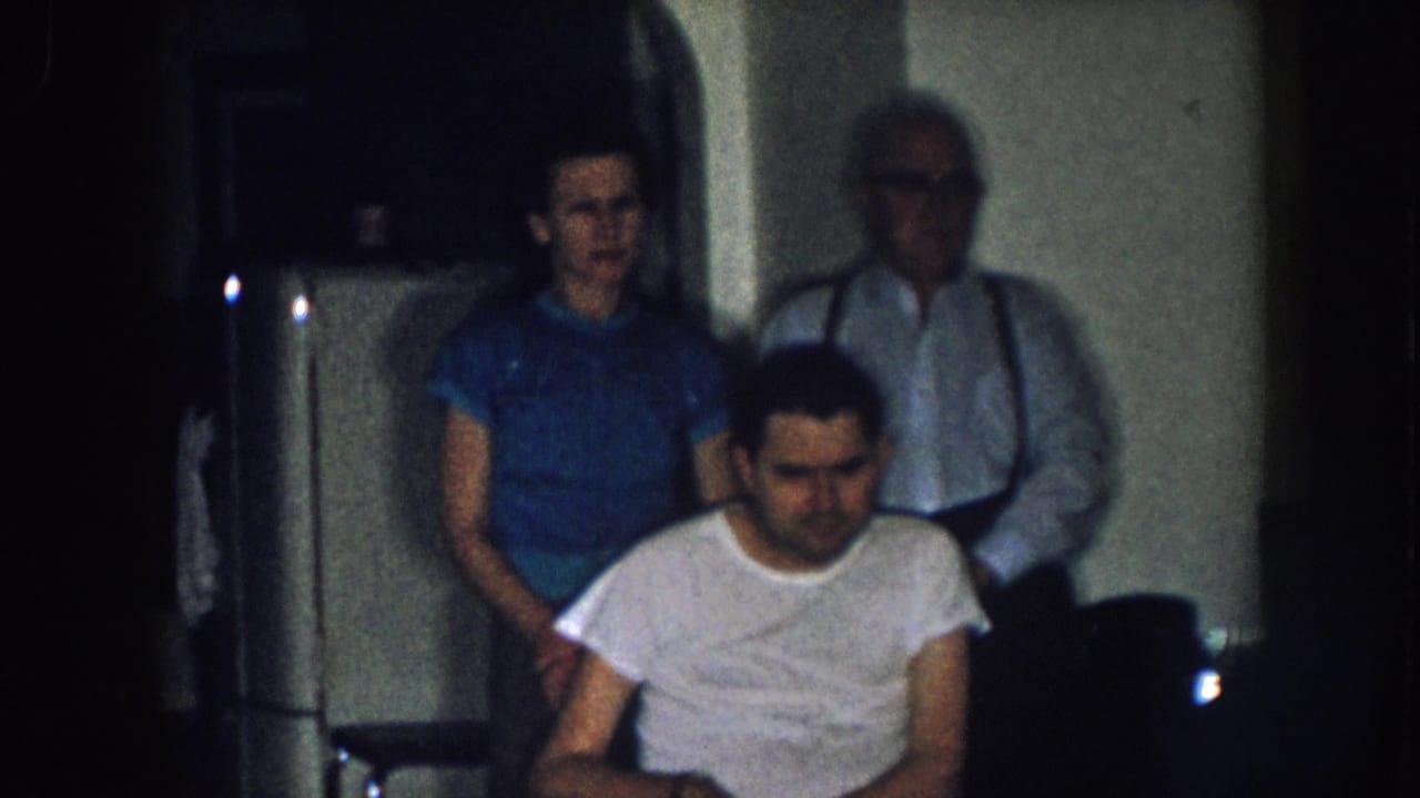Young adult man in wheelchair flanked by woman in blue shirt and older man with suspenders near a white appliance, domestic kitchen, Nebraska c. 1957 8mm home movie.