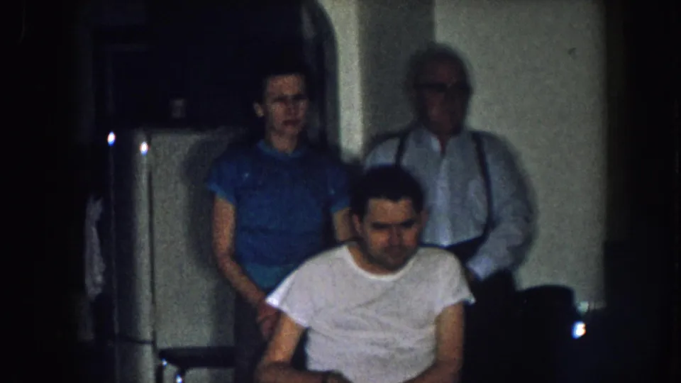 Young adult man in wheelchair flanked by woman in blue shirt and older man with suspenders near a white appliance, domestic kitchen, Nebraska c. 1957 8mm home movie.