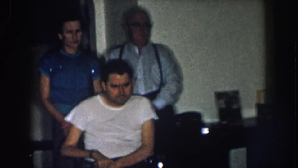 Three family members in domestic kitchen interior — young man in foreground, woman in blue shirt left, older man with suspenders right — Nebraska c. 1957 8mm home movie.