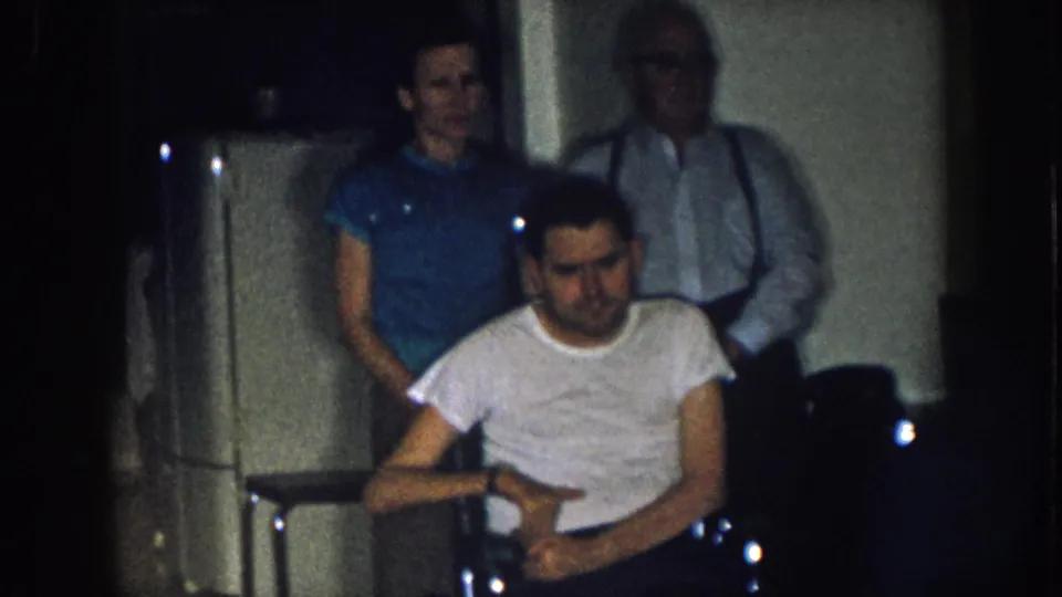 Young adult man in wheelchair with woman in blue shirt and older man with suspenders in a domestic kitchen interior, Nebraska c. 1957 8mm home movie.