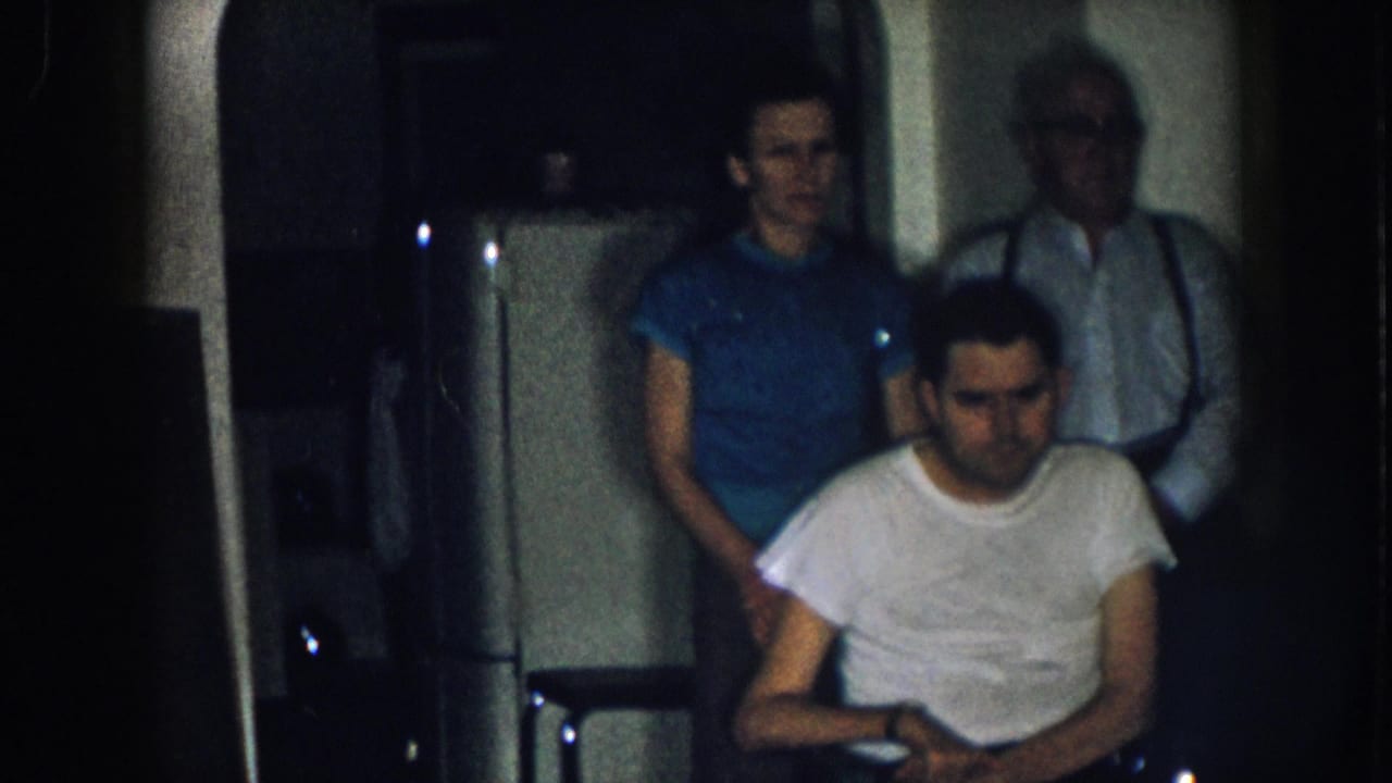 8mm home movie, circa 1957, Nebraska: seated man in white t-shirt with woman in blue top and older man in suspenders; light-colored appliance at left edge.