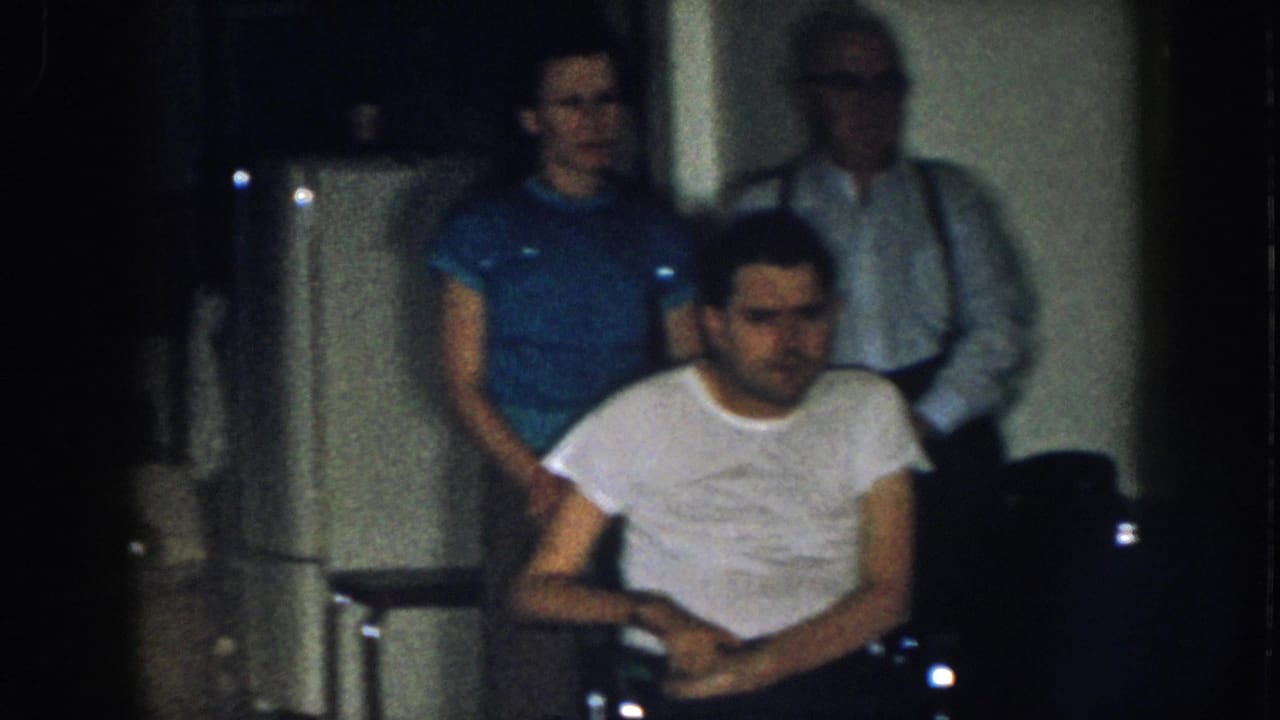 Same three adults in the domestic interior; the seated man appears to shift or fidget, woman in blue and older man in suspenders still visible behind him.
