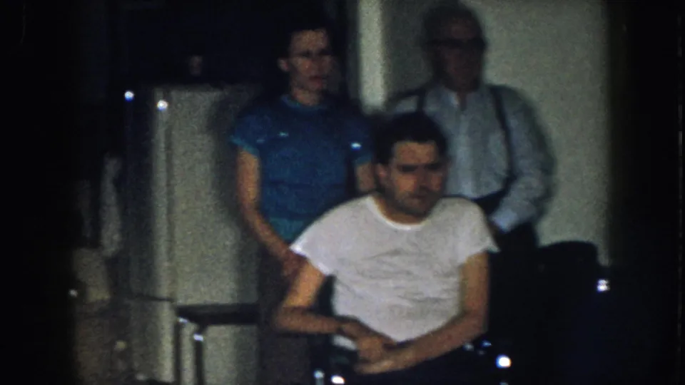 8mm home movie, circa 1957, Nebraska: seated man in white t-shirt appears to fidget while two adults stand behind him in a dim domestic interior.