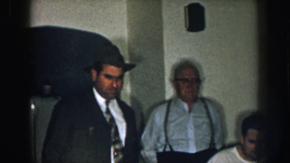 8mm home movie, circa 1957, Nebraska: suited man in fedora enters the scene, older man in suspenders behind him, arched domestic doorway visible in background.