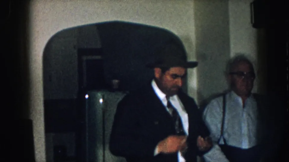 8mm home movie, circa 1957, Nebraska: man in dark suit and fedora foreground, older man in suspenders at right, arched domestic doorway and muted interior visible.