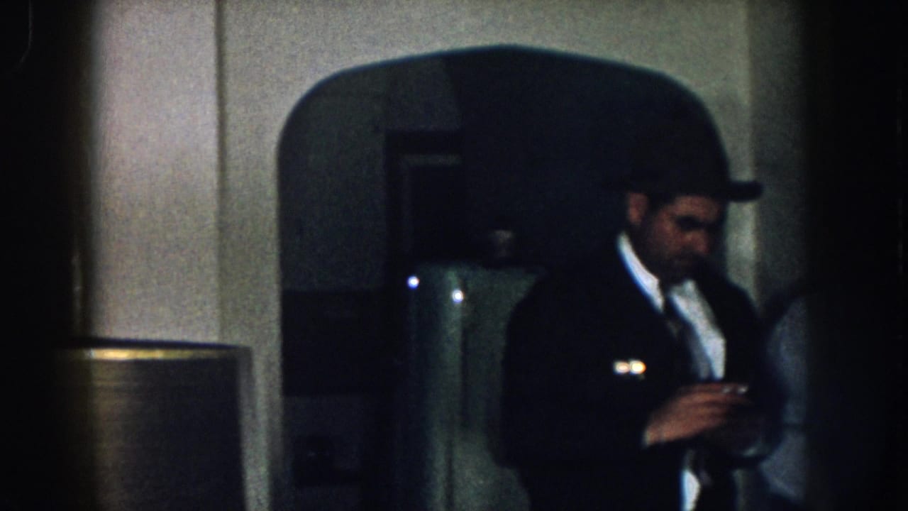 Two men in a dim 1950s Nebraska domestic interior; the foreground man wears a dark suit and fedora with a cigarette; a rounded-top refrigerator is partially visible behind them.