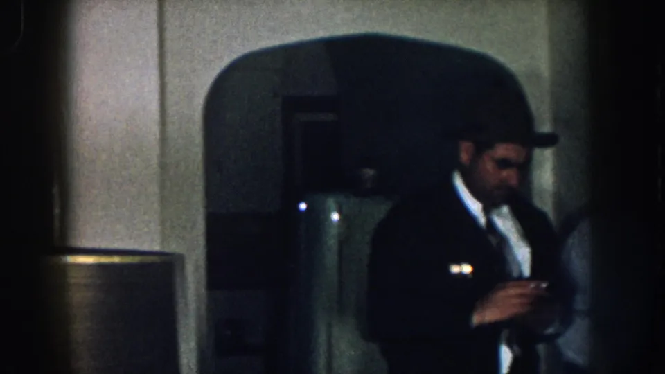Two men in a dim 1950s Nebraska domestic interior; the foreground man wears a dark suit and fedora with a cigarette; a rounded-top refrigerator is partially visible behind them.