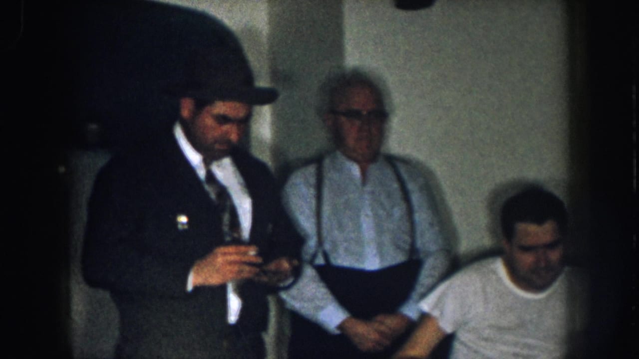 Three men in a dim indoor setting; one in a dark suit and fedora holds a cigarette, an older man in a light shirt and suspenders stands behind, and a man in a white t-shirt is seated at right.