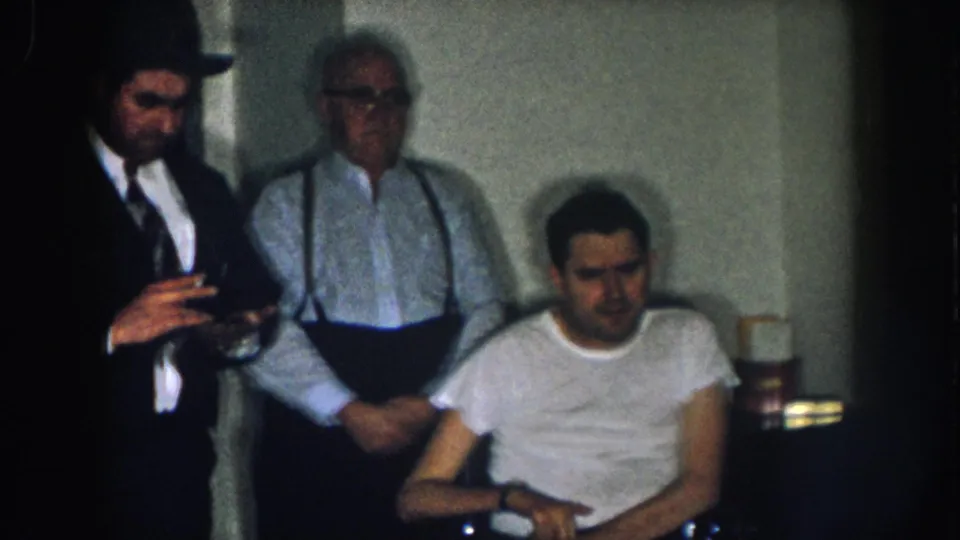 Candid 8mm frame from a 1950s Nebraska home: man in dark suit and fedora holds a cigarette, older man in suspenders and glasses stands nearby, younger man in white t-shirt sits in a manual wheelchair.