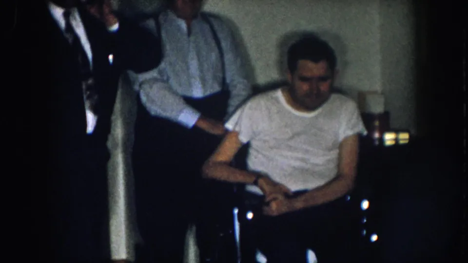Final frame of a 1950s 8mm Nebraska home movie: suited man with fedora at left, older man in suspenders at center, younger man in white t-shirt in a manual wheelchair at right, small object visible on a surface to the far right.