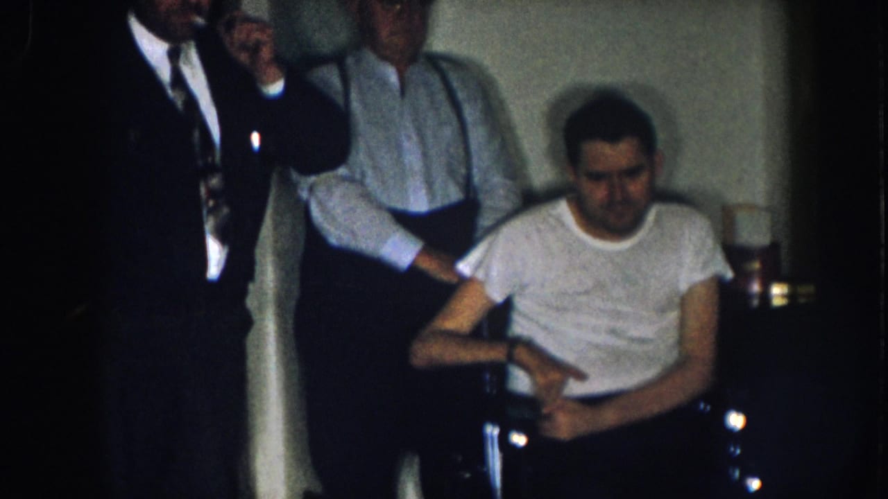Three men in a dim interior, Nebraska, 1957; one seated in a metal wheelchair in a white t-shirt, one in a dark suit and tie, one in a light shirt with suspenders.
