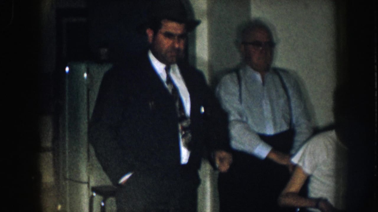 Man in dark suit and tie stands in foreground; two other men partially visible behind him in a dim interior.