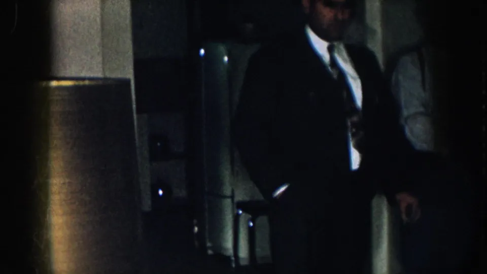 Close view of a man in a dark suit and tie looking downward in a dim interior; clothing style consistent with 1950s American dress.