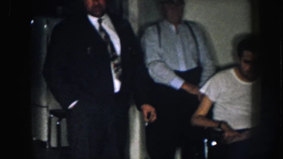 Three men in a dimly lit Nebraska interior, c. 1957; the wheelchair-seated man in a white t-shirt is at frame right while the suited man and the man in suspenders stand to his left.