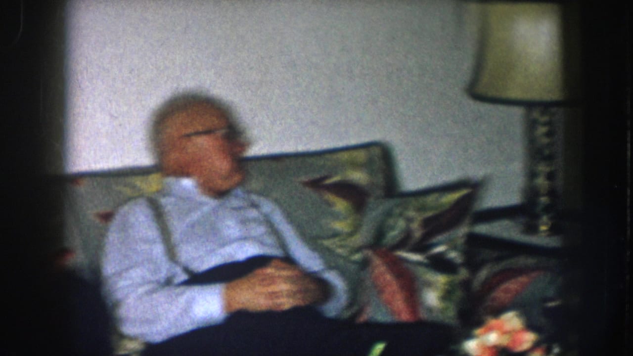 Older man with white hair, glasses, and suspenders relaxes on a floral-upholstered couch in a domestic interior, Nebraska, c. 1957.