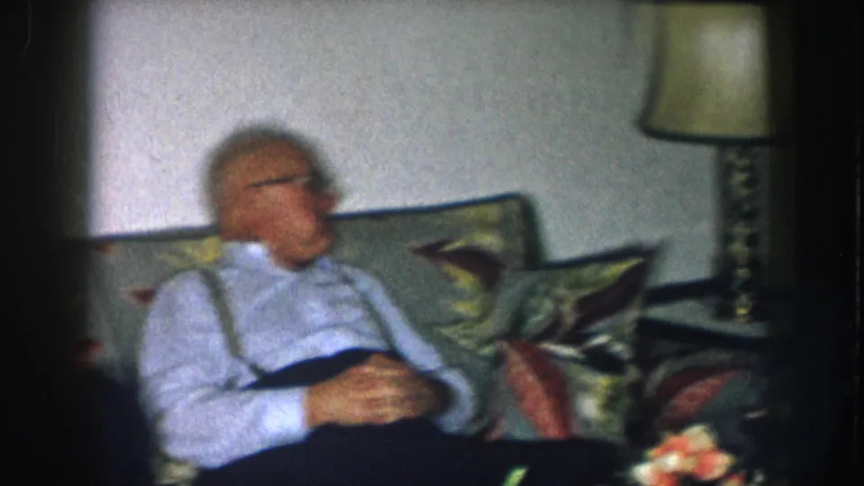 Older man with white hair, glasses, and suspenders relaxes on a floral-upholstered couch in a domestic interior, Nebraska, c. 1957.