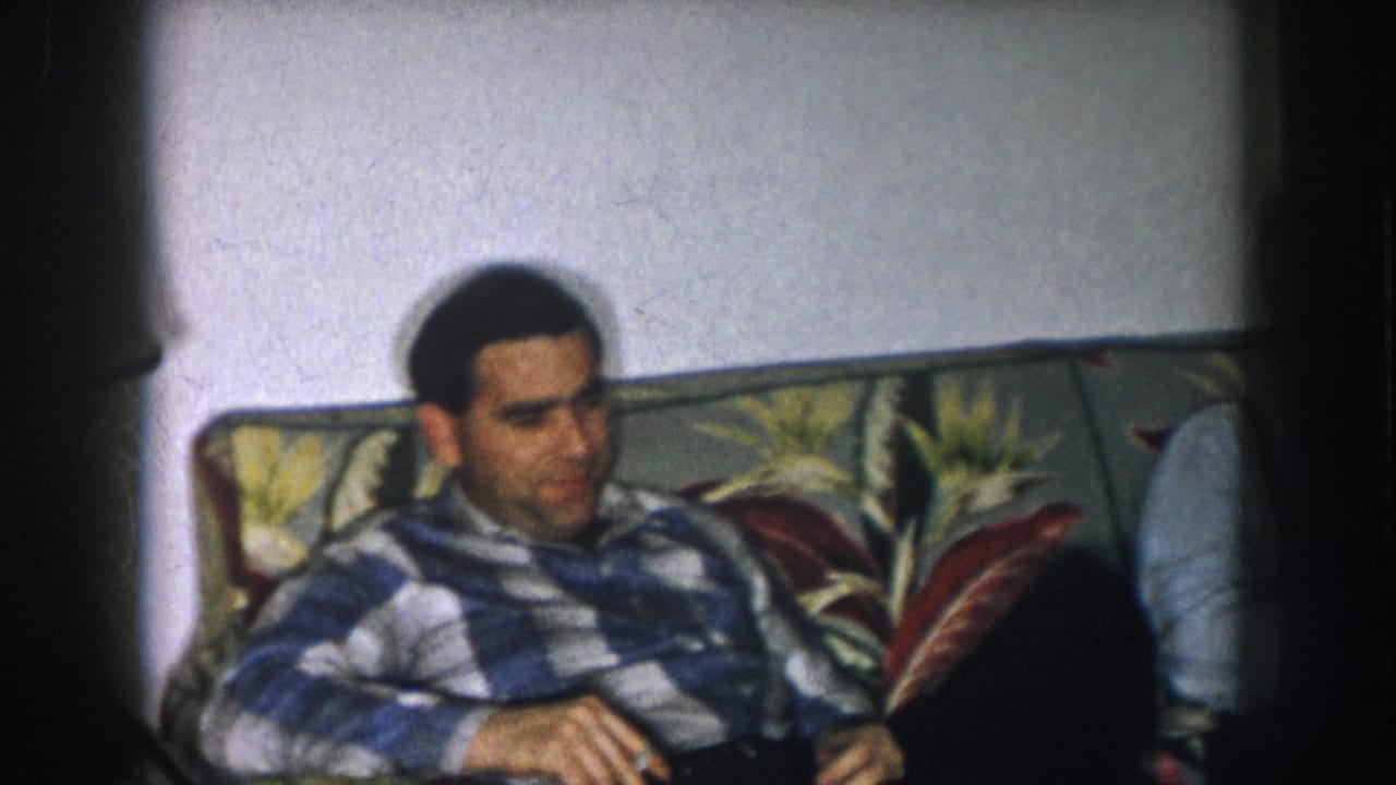 Young dark-haired man in blue-and-white plaid shirt seated on a bold floral-patterned couch, looking slightly downward.