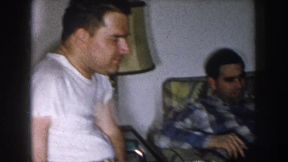 Two adult men — one in a white t-shirt, one in a plaid shirt — sit in a mid-century living room interior, Nebraska, c. 1957; both look away from the camera.