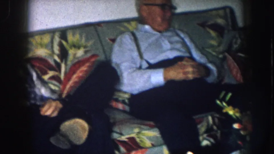 Older man with white hair, light blue shirt, suspenders, and glasses is seen again on the floral-patterned couch in a Nebraska domestic interior, c. 1957.