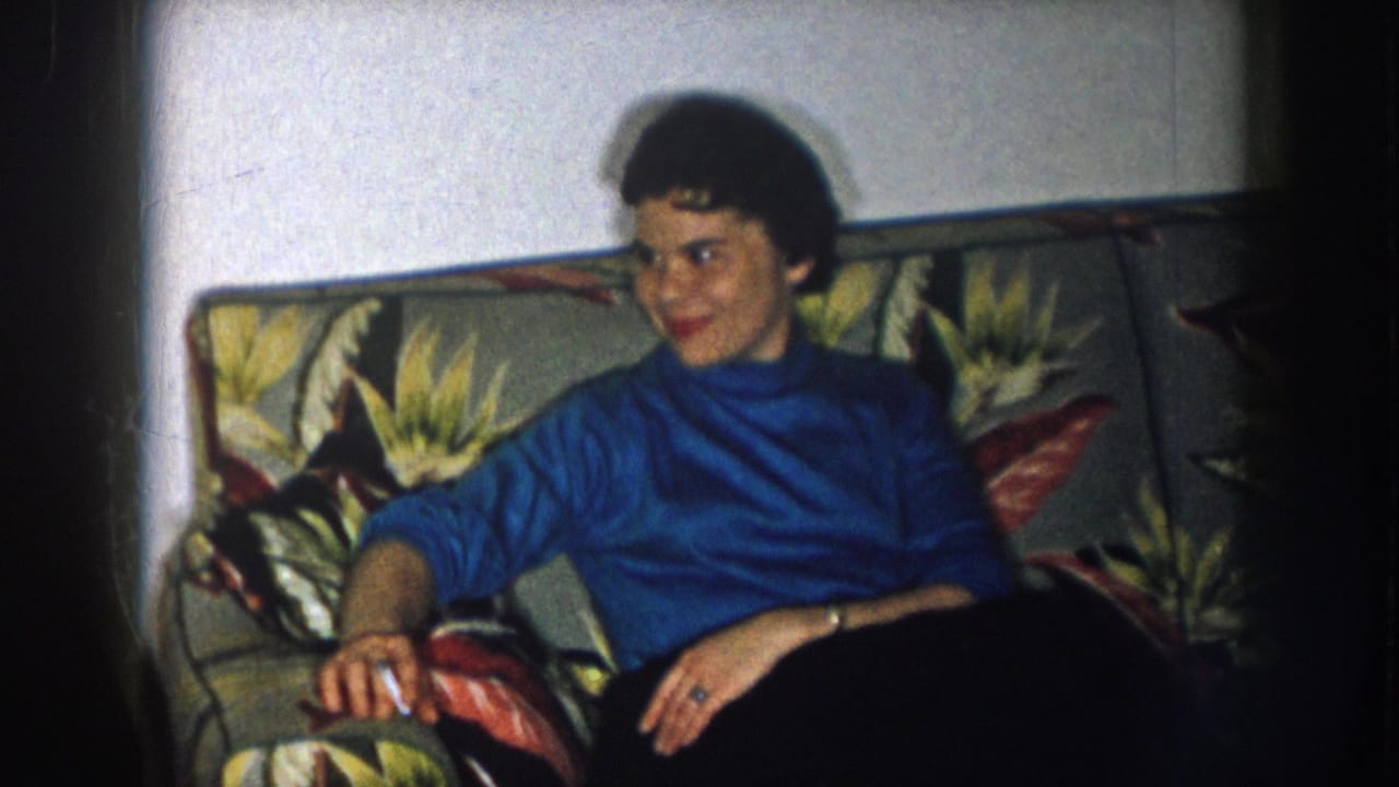 1957 Nebraska 8mm home movie; woman with short dark hair in blue turtleneck seated on tropical-print sofa, cigarette in hand, smiling toward camera.