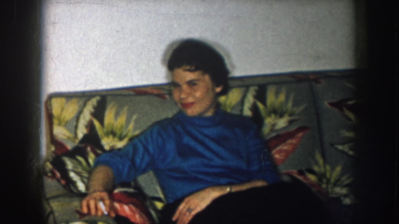 Woman in blue turtleneck on floral sofa, slight smile directed at camera, cigarette visible in right hand.