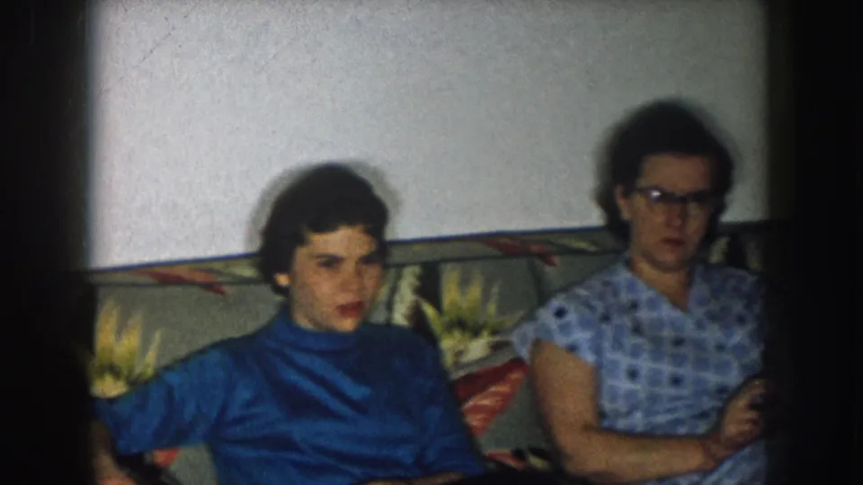 8mm home movie, Nebraska 1957; two women on a tropical-print sofa, left in blue turtleneck, right in checkered short-sleeve dress with dark-framed glasses.