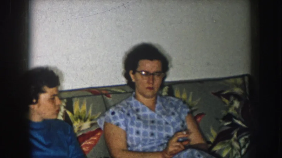 8mm home movie, Nebraska 1957; two women on floral-print sofa, second woman in blue-and-white checkered dress with glasses holding a cigarette.