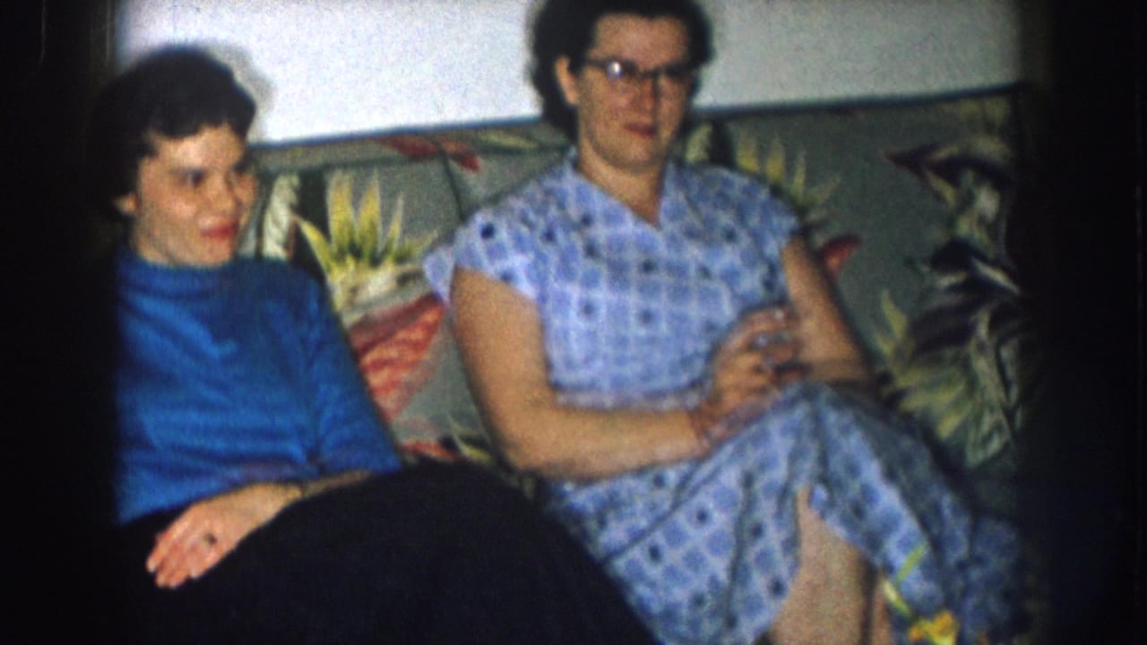 Two women on a floral couch in what appears to be a mid-1950s Nebraska living room — one in a blue turtleneck, one in a checkered dress and cat-eye glasses.